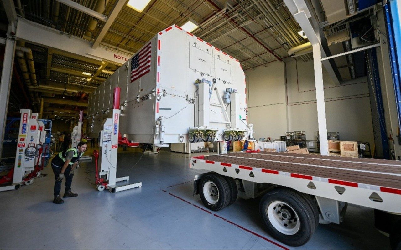  ​a male Lockheed Martin technician looks at a truck that is being loading with the GPS III SV07 satellite payload